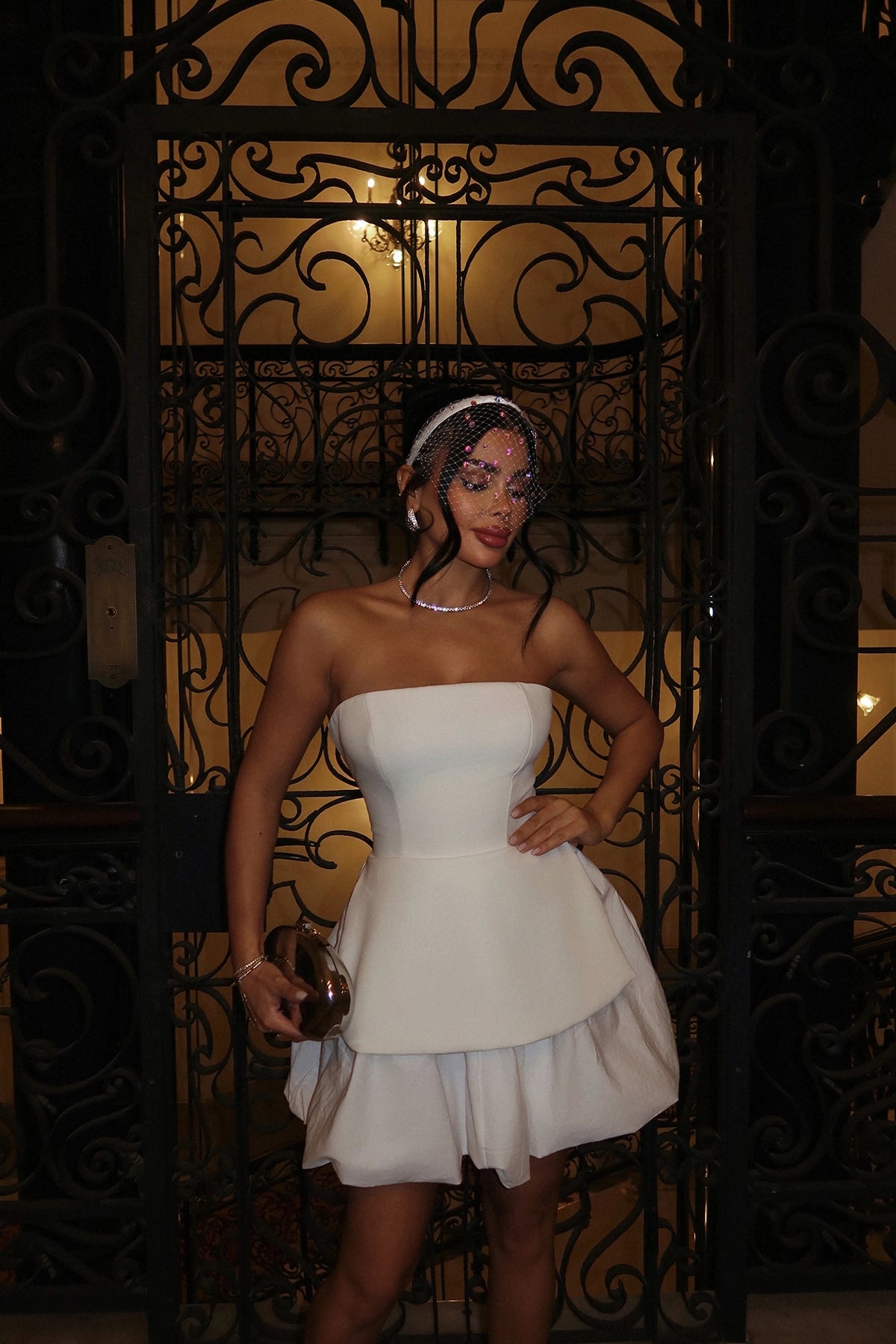 Woman in a strapless white peplum mini dress, metallic clutch in hand, posing in front of ornate black gates with chandelier lighting behind.