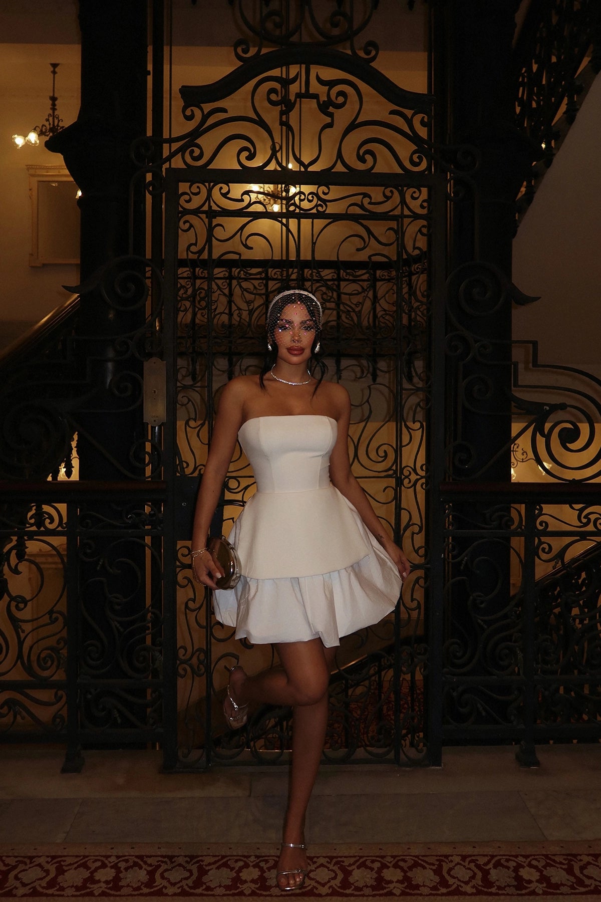 Woman posing in a strapless white peplum mini dress with layered skirt, holding a metallic clutch, standing against ornate black iron gates.