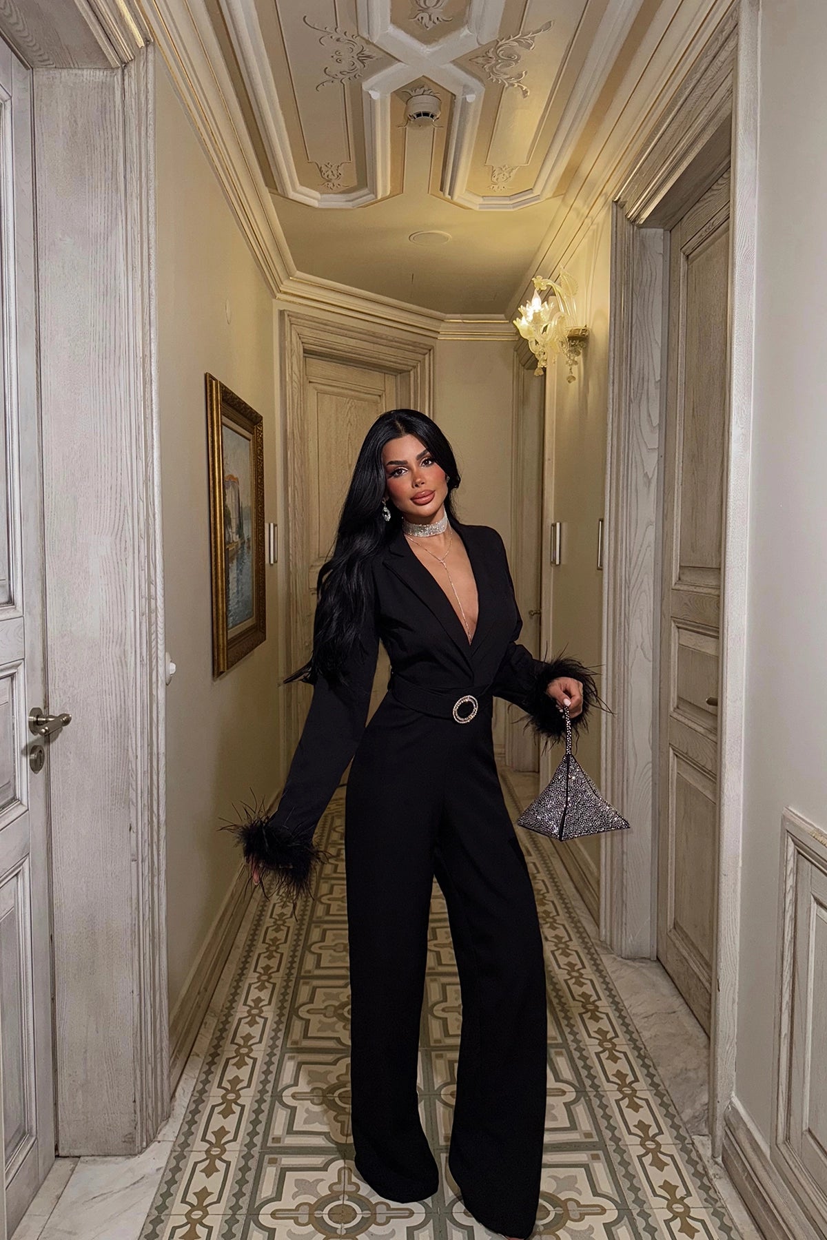 Woman wearing black belted jumpsuit with feather cuffs and deep V neckline.